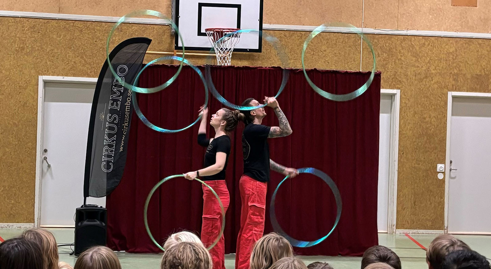 Två cirkusartister jonglerar med rockringar framför en skara med barn i en gymnastiksal.