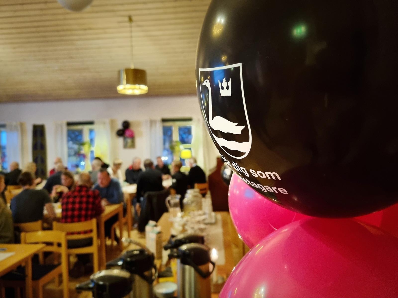 Svarta och rosa ballonger med kommunvapnet på sitter på en dörr. I bakgrunden syns människor som äter frukost. 