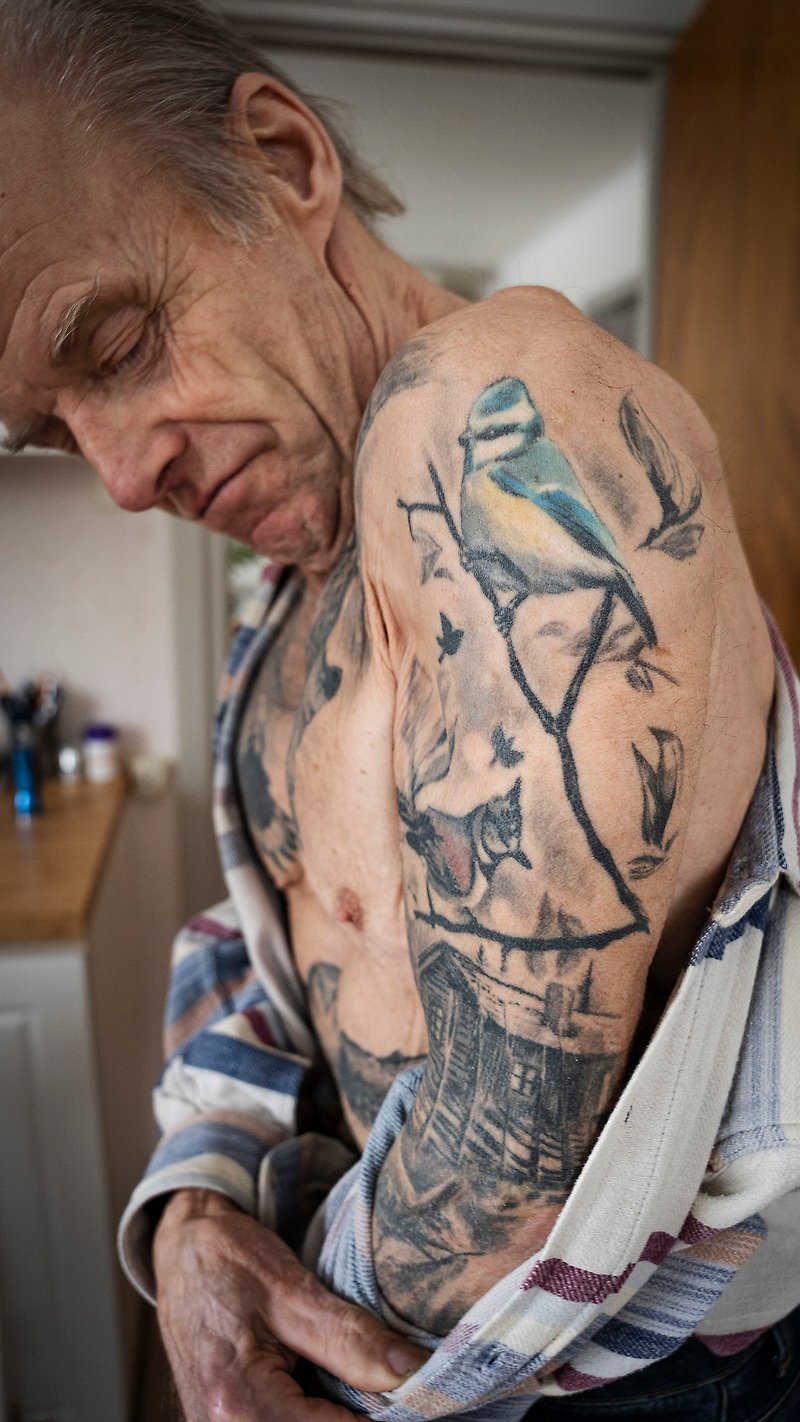 Alvar tar av sig skjortan och visar upp sin arm som är tatuerad med diverse fåglar och en stuga med snedhage.