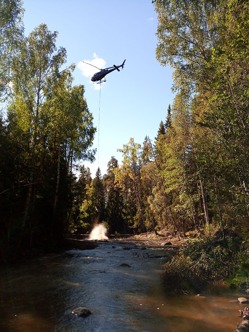 En helikopter släpper ner en behållare i en fors mitt i skogen så att det plaskar upp vatten.