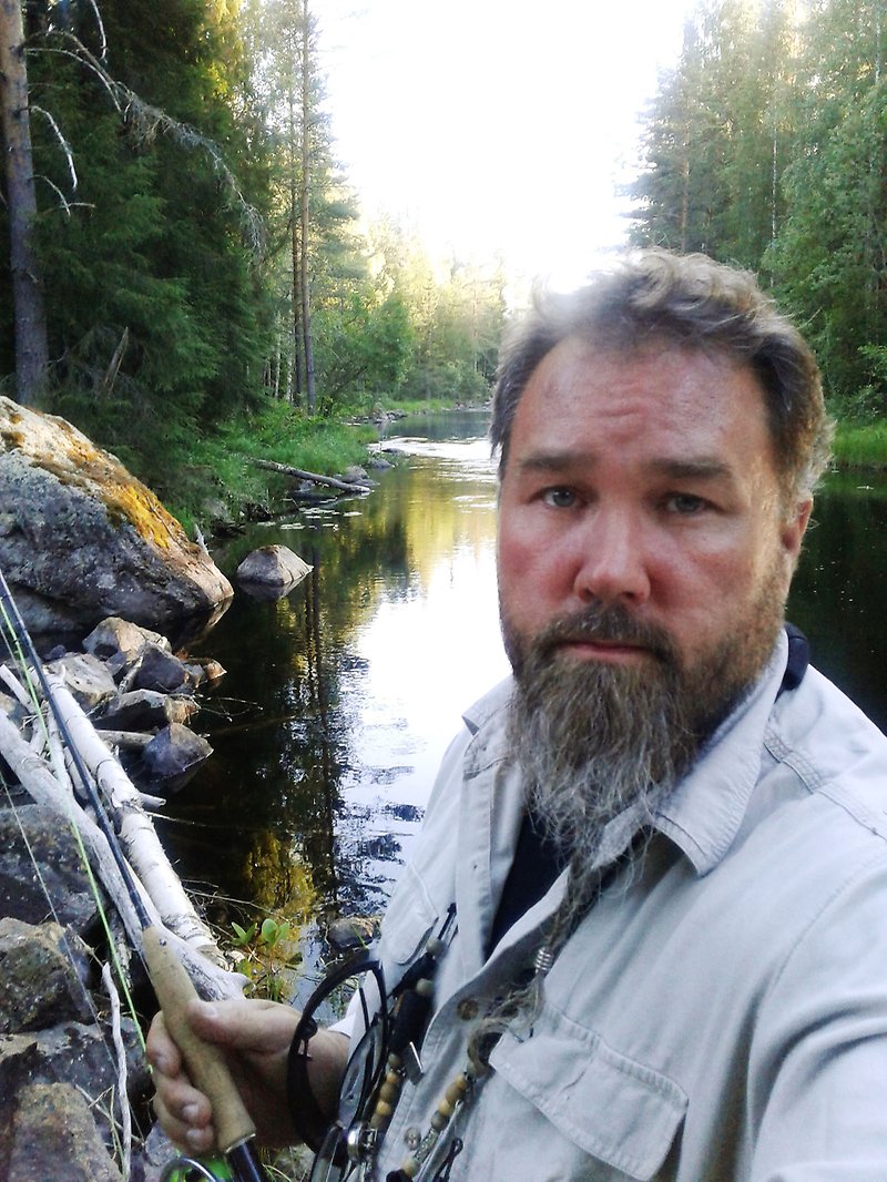 Porträtt av Christer Åsentorp som står vid en fors i skogen med ett fiskespö.