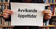 En skylt med texten "Avvikande öppettider" hålls upp av två händer framför en bokhylla.