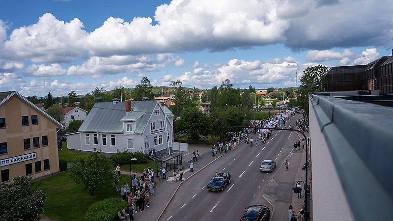 Ett tåg av studenter som går längs med långgatan i Edsbyn. Längst fram kör en fin bil. Därefter två personer med svenska fanan i händerna.