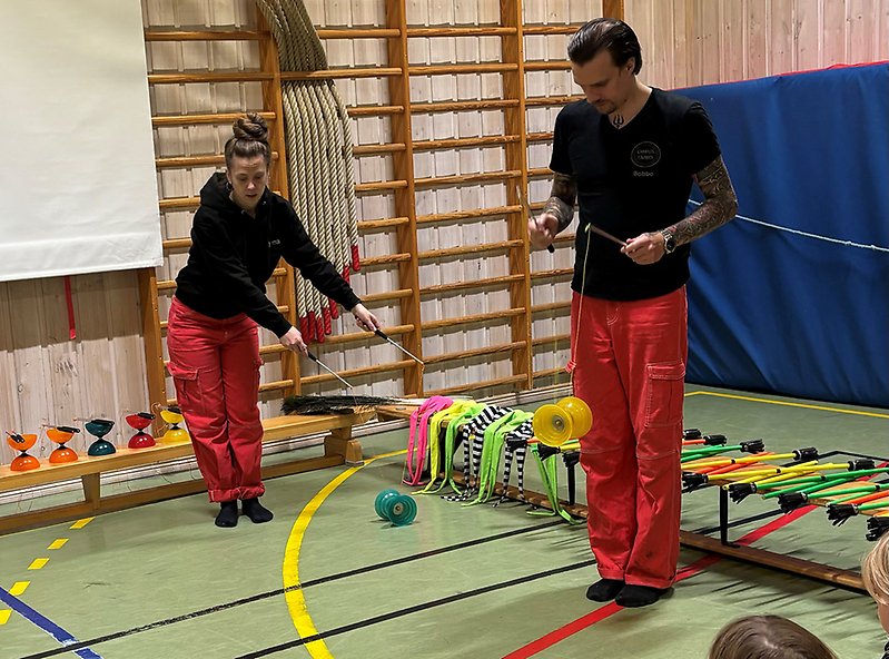 Två cirkusartister snurrar med diabolo framför en skara barn i en gymnastiksal.