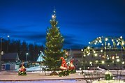 En julgran med tända lampor i står tillsammans med tre tomtar med lampor på mitt i en rondell.